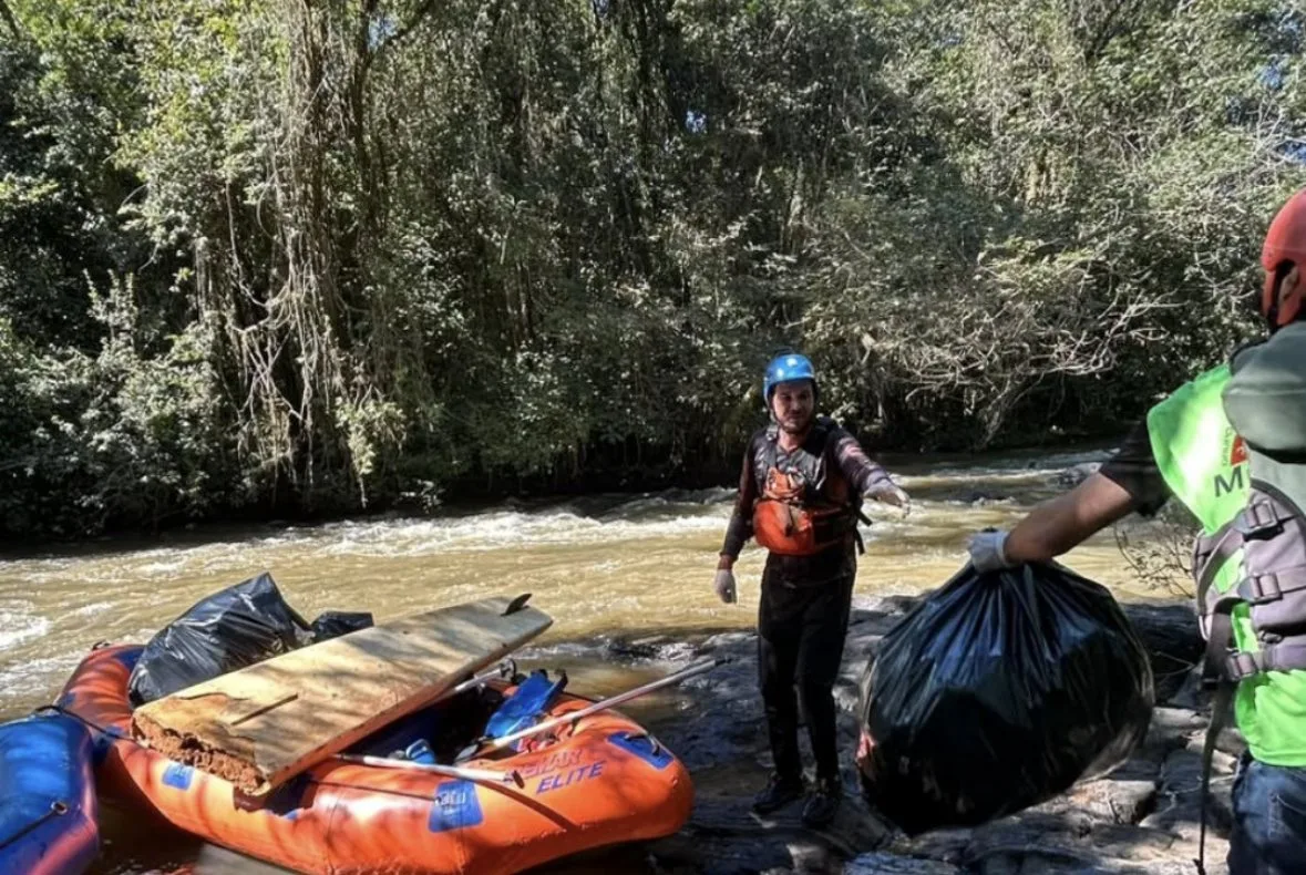 Voluntários com equipamentos de proteção retiram sacos de lixo do rio, ao lado de um bote.